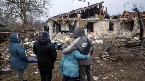 Four people in winter jackets look at ruined house.