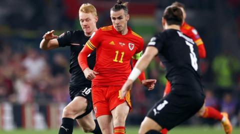Gareth bale in Wales kit dribbling between two austrian players in black kit. Gareth has an intent look and has his usual Bale bun ton knot hairstyle