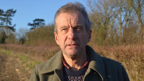 Michael Dunk is standing in a field with a hedgerow and trees in the background. He is wearing a dark green coat and a patterned jumper. He is not looking directly at the camera but is looking to the left of the image.