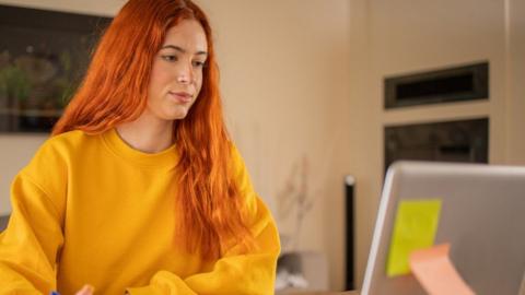 A student with long red hair and a bright yellow jumper looks at her laptop screen.