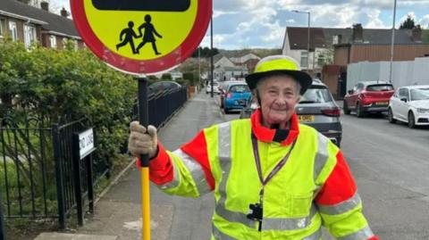 An elderly woman in a crossing warden's hi-viz jacket and hat, holding the lollipop sign. She stands on a pavement next to a road with lots of cars parked on it.