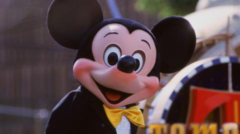 The Disney character Mickey Mouse in a Disney World theme park, smiling widely and wearing a gold bow-tie.