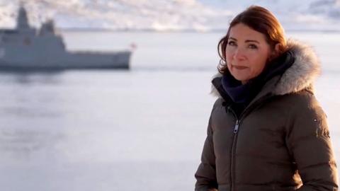 Katya Adler in a coat by the sea with a boat in the background.