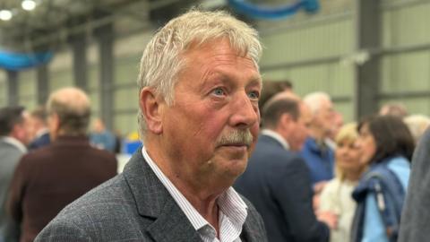 A photo of Sammy Wilson at an election count in 2024. He has short grey hair and a grey moustache. He is wearing a grey suit with a subtle checked pattern and an open-necked white and grey striped shirt. Crowds of people are talking in groups in the background.