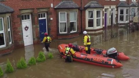 Aerial view of three rescue workers in yellow and black drysuits and red life jackets and helmets approaching a house in a terrace all of which are flooded. One of the workers is standing in an inflatable boat, another is up to the top of their thighs in the water and a third is wading through water at knee height towards a white front door.