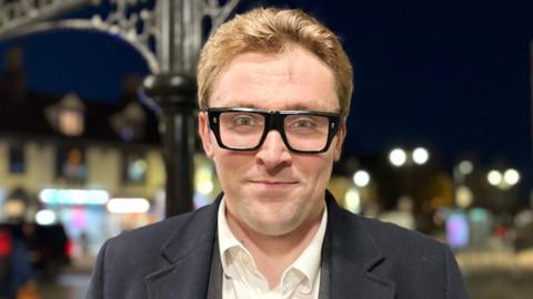 Alexander Curtis with short ginger hair and black-framed glasses. He is wearing a white shirt, grey jacket and dark grey coat. He is standing in a town centre street at night with lights in the background. There is a two-storey building behind him.
