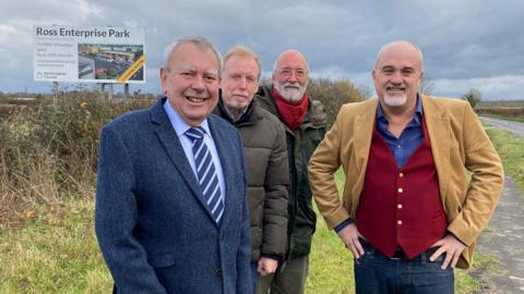 Four men standing outside in a field, with a large sign behind them that says 'Ross Enterprise Park'.