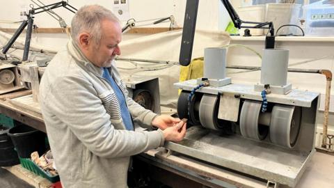 A man is standing in front of a grind machine. He is holding a stone in front of the machine. The machine has four circular conveyor belt wheels which is used to grind stones.