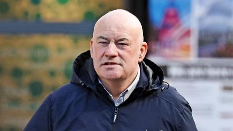 A bald man wearing a black zip-up rain coat over a pale shirt. The background is out of focus.