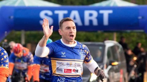 Kevin Sinfield wearing a blue and white race jersey with sponsor logos raises one hand near the starting line. The background features a large blue inflatable arch with the word ‘START’ in bold white letters.