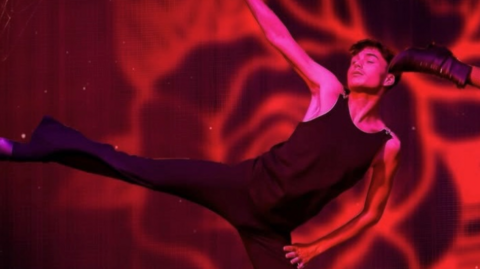 Finn on stage in a leotard and with his arms outstretched. His right leg is outstretched and he is looking off to the side against a red swirling background.