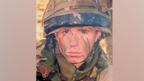 A head and shoulders image of Simon Miller wearing army camouflage clothing and helmet, with brown and green stripes on his face. He has blue eyes and is looking directly at the camera.