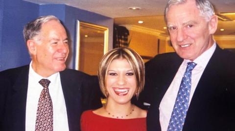 A smiling woman in her 20s with bobbed blond hair and wearing a red top, stands between two older men with white hair who are wearing suits and ties. All three of them are smiling.