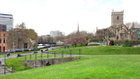 Castle Park in Bristol. A church can be seen on the far right with a green lawn in front of it with walkways running through it. The river is visible on the left. It's a grey day and city buildings can be seen in the background.