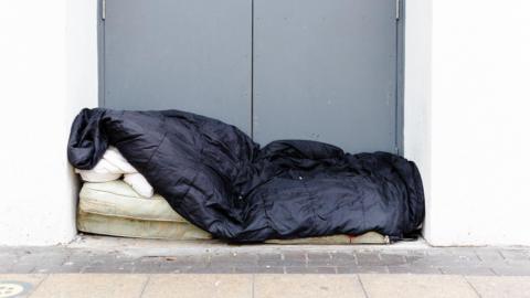 A mattress with a black sleeping bag over it in a doorway on the pavement.