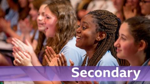 Teenage girls sat in an audience applauding. They are wearing blue school uniforms. Text reads, 'Secondary'.