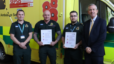 Three people are in ambulance uniform and one is in a suit. They are stood in front of an ambulance which has a poppy design on the side. The two people in the middle are holding white certificates.