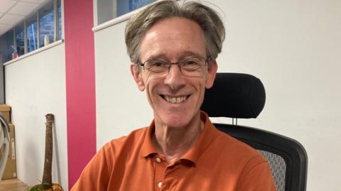 A mid shot of Marko Kennedy. He has short grey hair and is wearing glasses, and an orange polo shirt.