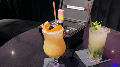 Two cocktails - one yellow, another looking suspiciously like a mojito, on a table in front of a black box containing a machine which contains a vape