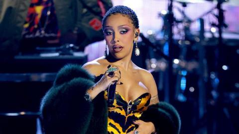 Doja Cat is performing on stage. She is singing into a mic which she is holding in front of her with her left hand. She is wearing a black and orange tiger print bustier top, which has a black and silver piece of hardware in between the bra cups. On top of that is a black fur coat, which is across her arms and not covering her shoulders. Doja Cat is wearing her natural hair and heavy, glittery eye make-up. She has dangly earrings with large white gemstones and an ear piece in one ear. On her fingers are various large rings and she has long black stiletto-shaped nails.