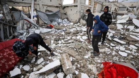 Three young people watch a fourth who is sifting through the rubble of a residential area