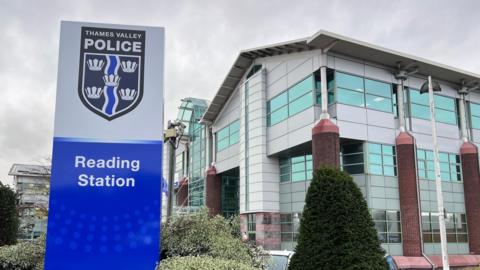 A general view picture of Reading Station, with a sign saying "Reading Station" and Thames Valley Police insignia above that. Behind it stands a modern building.