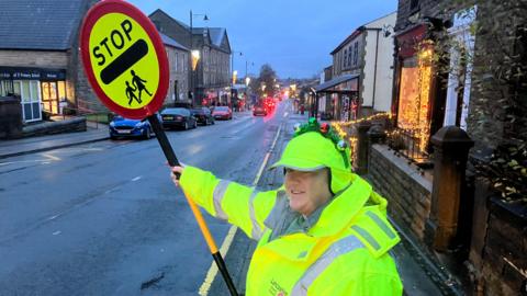 A woman in a yellow high-vis coat and festive headband holds a stop sign over a road. Behind her cars can be seen queuing in the background.