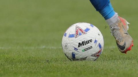 A National League matchball with HUFC on it being kicked by a white boot with a blue sock