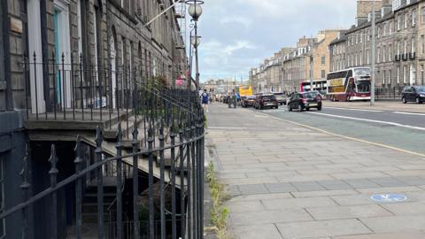 Railings marking the area of a basement. There is a pavement with steps rising up to a green doorway above. there are cars on the street.