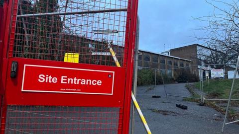 A red gate on the left hand side with a sign saying Site Entrance and more metal fencing in front of the derelict police station site with boarded up windows.
