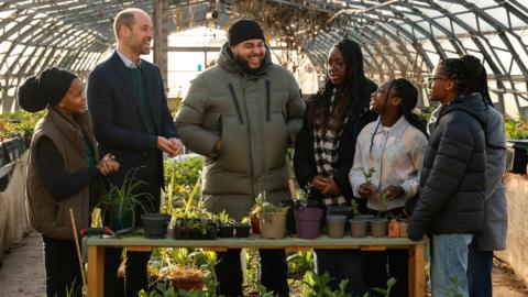 Four teenage girls can be seen stood around a table which has several potted plants on it inside a greenhouse. There is also a man stood in the middle laughing and next to him is Prince William who can be seen looking at the girls and smiling. A woman is stood next to him also smiling.
