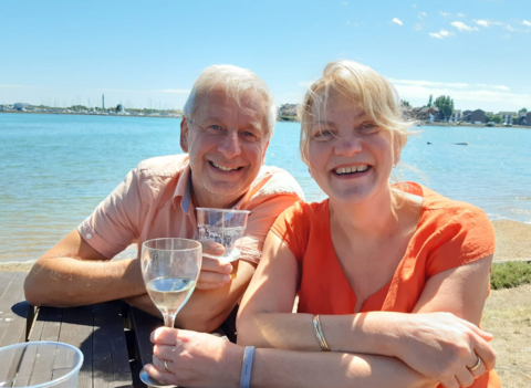 Chris and Ruth Stone-Houghton sit on a picnic bench, smiling into the camera. They are holding alcoholic drinks, and behind them is a body of water