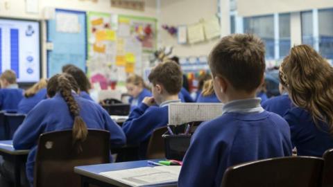 Children in blue uniforms sitting in chairs in a classroom, a mix of boys and girls seen from behind