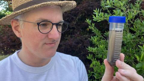 James Gallagher wearing a white T-shirt and summer hat, looking closely at a vial containing pond-water