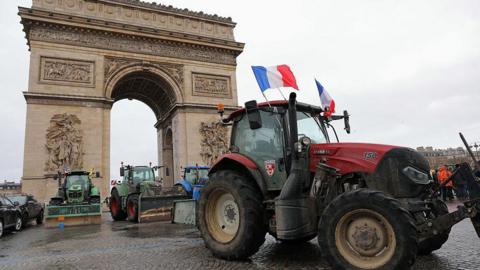 Several tractors, including a red tractor flying French flags, are parked in front of the Arc de Triomphe in Paris.