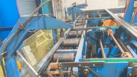 A blue machine with open cogs and chains. There are various metal components which are unscreened. The machine has a row of rollers and other workings. There are unpainted metal shutters in the background.