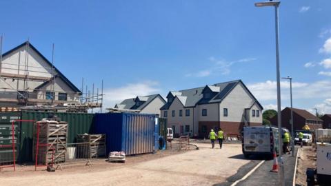 A new build housing estate nearing completion on a sunny day. One home still has scaffolding on it. Vans and other larger vehicles are parked around the site. People wearing hard hats and high-vis jackets can be seen.