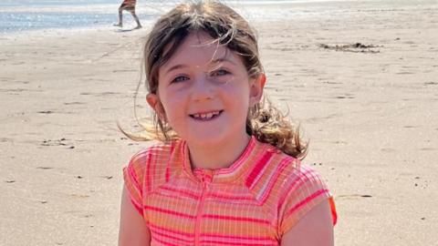 A young girl in a swimsuit sits at the beach. Her brown hair is tied into a ponytail and she smiles at the camera.