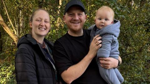 A man and woman are standing together in an outdoor setting. The woman on the left is wearing a dark jacket and is smiling a the camera. The man is wearing a black shirt and cap is also smiling and is holding a baby. The baby is dressed in a light-coluored hooded baby grow. Behind them are trees and foliage.