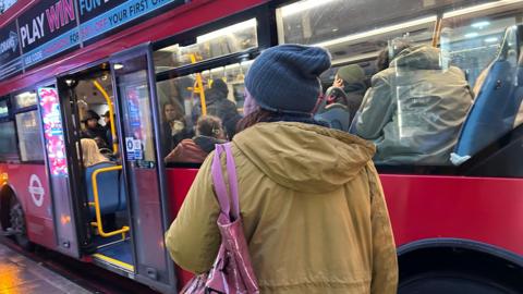 A woman in a coat stands facing away from the camera next to a London bus