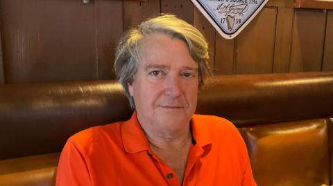 The image shows a man wearing an orange top. He is looking directly at the camera. He is sitting on a brown padded seat and there is a wooden wall behind him.