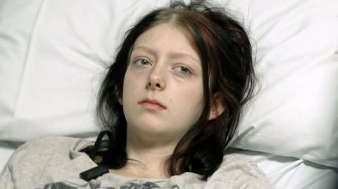 Paige who is 19 is lying on a hospital bed resting her head on a pillow. She looks ill. She has dark hair and is wearing a T-shirt