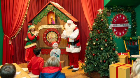 A grotto scene with a very large cuckoo-type clock and Santa and an elf standing by with children taking part and watching. There is a large Christmas tree to the right and a sign saying 'Santa O'Clock' in red and white