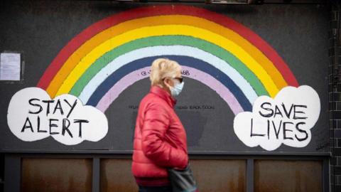 A woman wearing a face mask, sunglasses and a red coat walks past a rainbow mural on a wall which reads "Stay alert save lives" during the Covid pandemic.