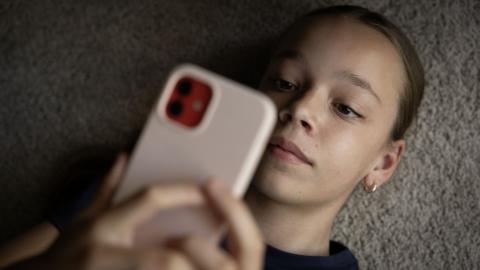 A girl lies on grey carpet and looks at her phone which is in a pink case