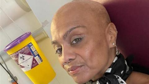 A woman looking straight at the camera while sitting in a red hospital chair. She is in front of a yellow and purple hazardous waste bin.