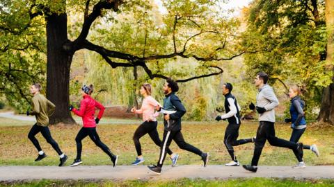 A group of seven runners run through a park. They are all wearing black bottoms and colourful jackets.