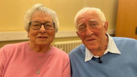 Wendy, wearing a pink top, and Ken, in a blue jumper, are sitting in a police station looking at the camera during their interview.