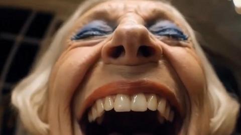 An extreme close up of the face of an elderly lady with blue eye make up on and grey hair laughing with her eyes closed and mouth open wide.