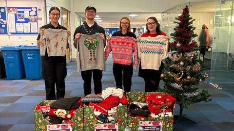 Four Hull City Council staff standing in an office next to a Christmas tree hold up some of the donated sweaters in front of Christmas decorated boxes holding more jumpers